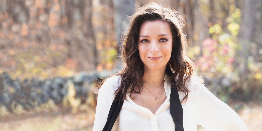 Yulianna Avdeeva wears a white blouse with a black scarf flowing around her next with her hand on her hip.
