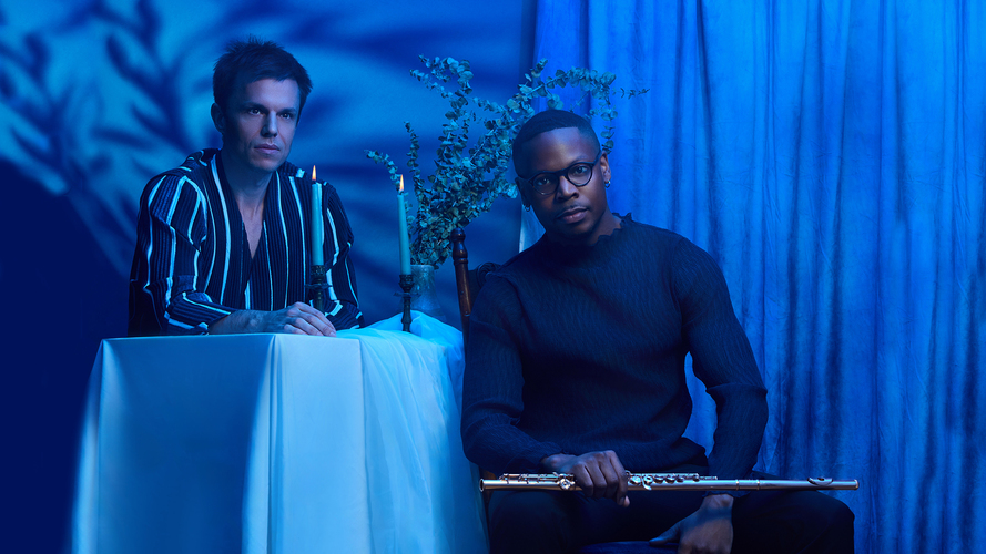 Brandon Patrick George sits and Bryan Wagorn stands at a table under blue light.