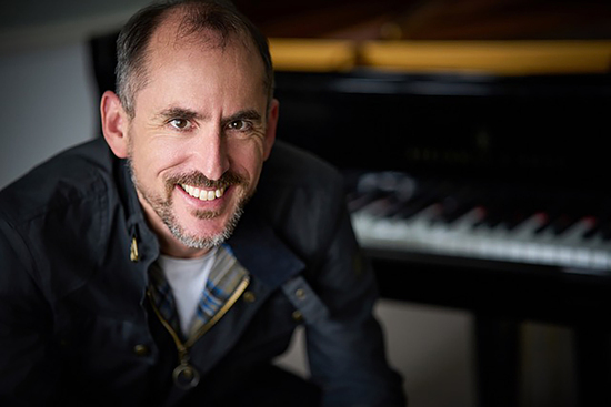 Danny Driver smiles in front of a piano.