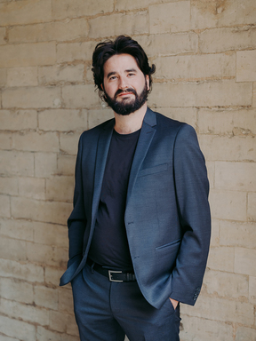 Julien Brocal wears a blue suit with hands in his pockets and standing against a white brick wall.