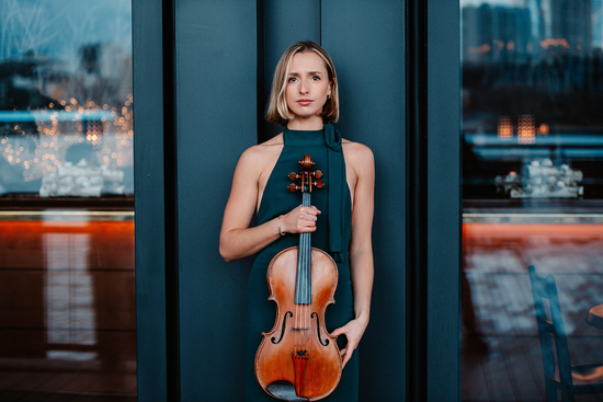 Natalie Loughran stands wearing a green jumper holding her viola.